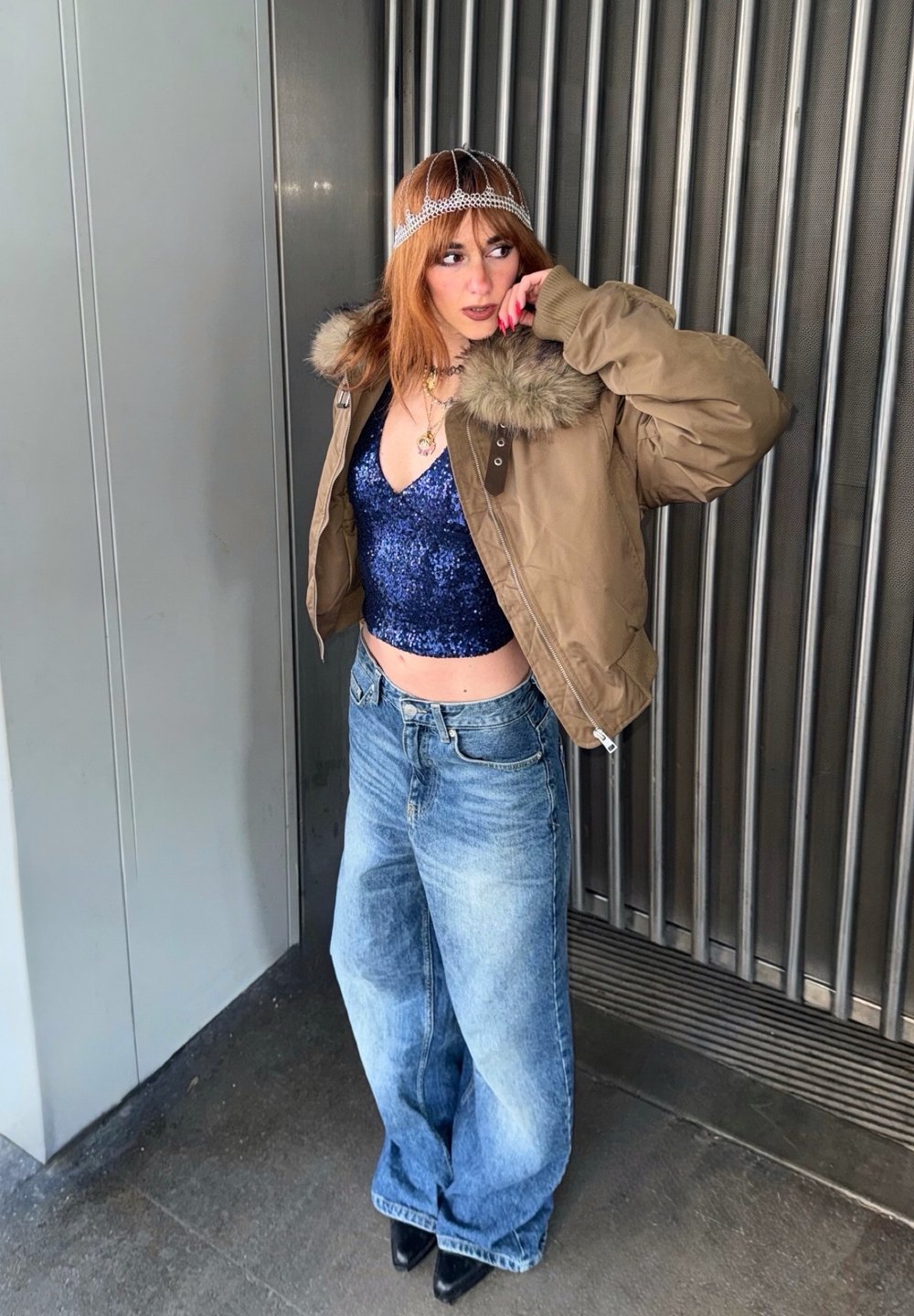 Personne aux cheveux roux portant une veste à col en fourrure marron sur un haut à sequins bleu, un jean à jambes larges, des chaussures noires et un ornement de tête décoratif, debout à l'intérieur.