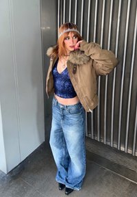 Personne aux cheveux roux portant une veste à col en fourrure marron sur un haut à sequins bleu, un jean à jambes larges, des chaussures noires et un ornement de tête décoratif, debout à l'intérieur.