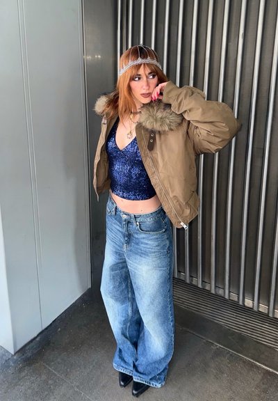 Persona con cabello rojo que lleva una chaqueta con cuello de piel marrón sobre una blusa azul con lentejuelas, jeans de pierna ancha, zapatos negros y un adorno decorativo en la cabeza, de pie en el interior.