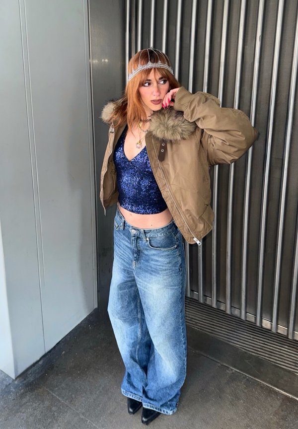 Person mit roten Haaren, der eine braune Jacke mit Pelzkragen über einem blauen Paillettenoberteil, weit geschnittene Jeans, schwarze Schuhe und ein dekoratives Kopfschmuck trägt, steht drinnen.
