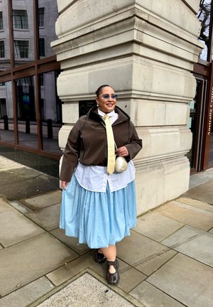 Persona con gafas de sol, chaqueta marrón, corbata amarilla, camisa blanca, falda azul clara y zapatos negros de pie junto a la esquina de un edificio de piedra en una acera.