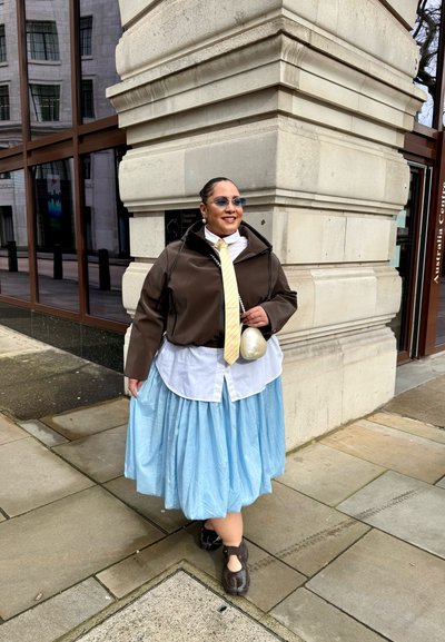 Persona con gafas de sol, chaqueta marrón, corbata amarilla, camisa blanca, falda azul clara y zapatos negros de pie junto a la esquina de un edificio de piedra en una acera.