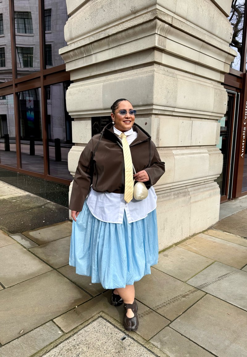 Personne portant des lunettes de soleil, une veste marron, une cravate jaune, une chemise blanche, une jupe bleu clair et des chaussures noires, debout au coin d'un bâtiment en pierre sur un trottoir.