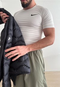 Homme portant un t-shirt blanc Nike et un pantalon vert, tenant une veste noire matelassée avec un logo blanc dans un cadre intérieur simple.