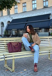 Μπεζ υπερμεγέθη παλτό, πράσινο πουκάμισο και μπλε τζιν. Καφέ τσάντα πάνω σε παγκάκι. Το μοντέλο κρατάει μια κούπα. Κόκκινα μποτάκια με λεπτομέρεια αλυσίδας, πετρόστρωτο έδαφος.