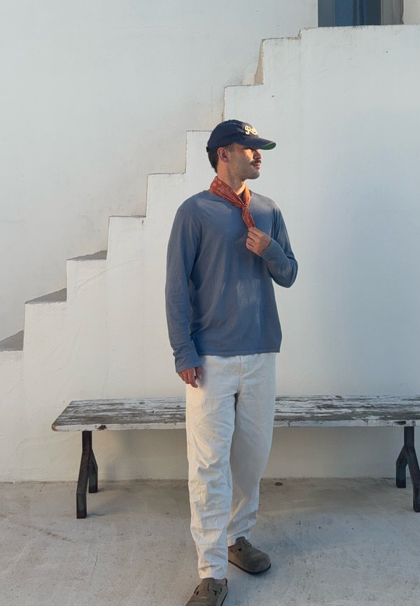Hombre con camisa azul de manga larga, pantalones blancos, gorra y bufanda naranja, de pie junto a una escalera blanca y un banco de madera contra una pared blanca.