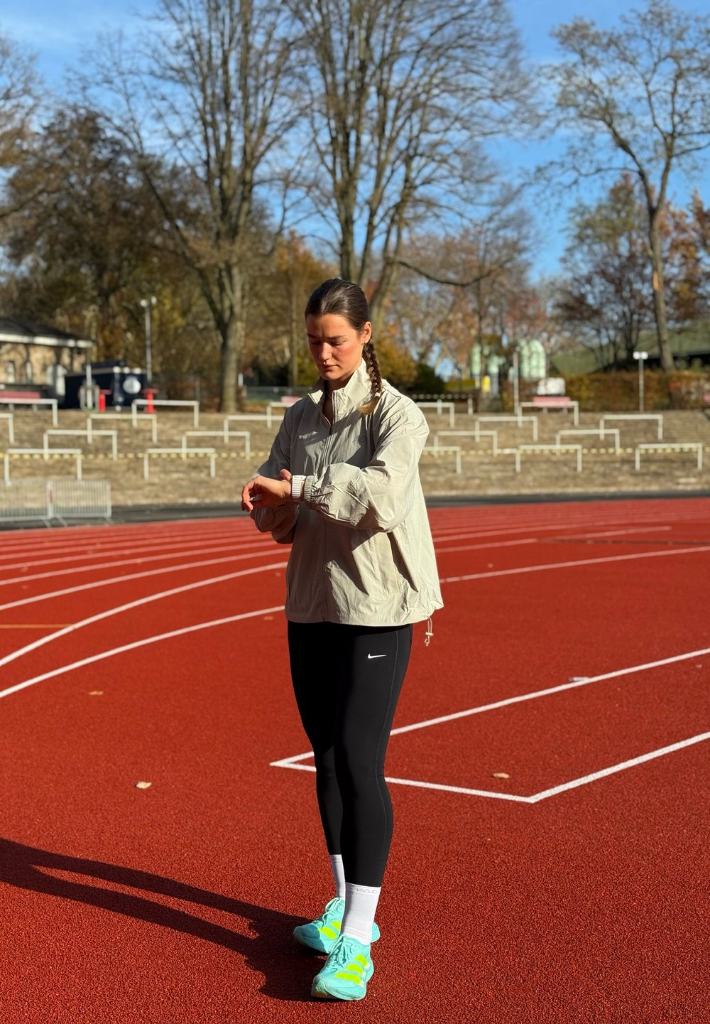 Ljusbeige vindjacka, svarta leggings, turkos löparskor med vita detaljer, står på en röd löparbana och tittar på en klocka.