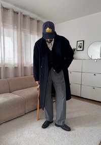 Homme portant un pardessus sombre, un pantalon gris, une cravate rayée et une casquette marine, debout dans un salon avec un canapé beige et une unité de rangement en métal blanc.
