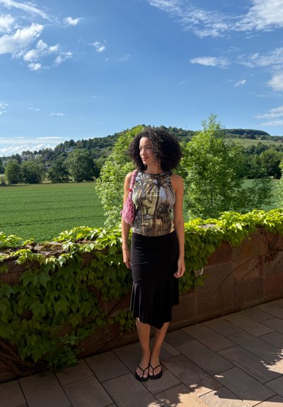 Mujer con pelo rizado que lleva una blusa sin mangas con estampado y una falda negra, de pie en un patio junto a un muro de piedra cubierto de enredaderas con vegetación detrás.