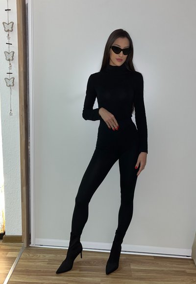 Mono negro ajustado con mangas largas y cuello alto, combinado con tacones negros de punta. La modelo lleva puestas gafas de sol negras elegantes.