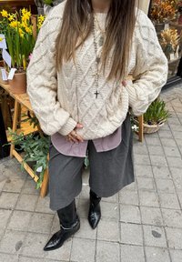 Maglione a coste a treccia crema indossato sopra una camicia a scacchi, abbinato a pantaloni grigi a gamba larga e stivali neri a punta, completato da collane.
