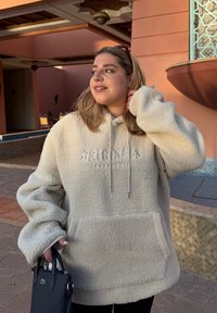 Lys beige fleece hettegenser med frontlomme og snor, med "ORIGINALS" preget på brystet. Bæres med en svart håndveske.