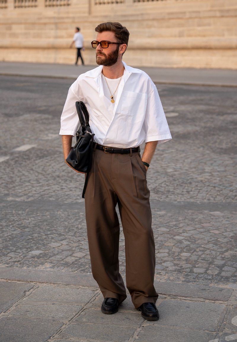 Homme portant une chemise blanche, un pantalon marron à jambes larges, des lunettes de soleil et des chaussures noires, portant un sac noir dans une rue de la ville.