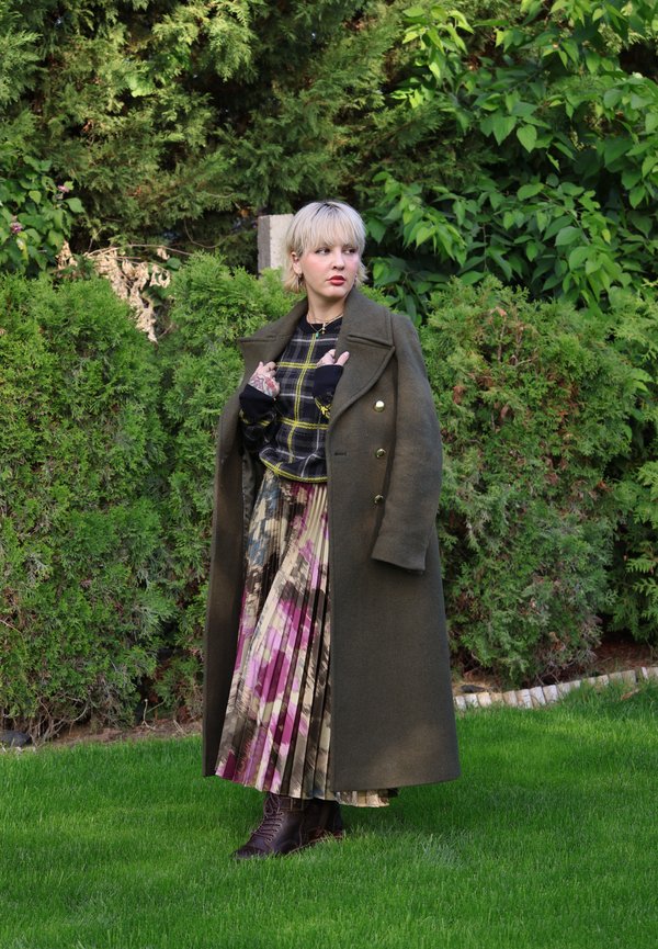 Una mujer rubia con un abrigo largo color oliva, blusa a cuadros, falda estampada y botas está de pie sobre el césped frente a arbustos densos del jardín.