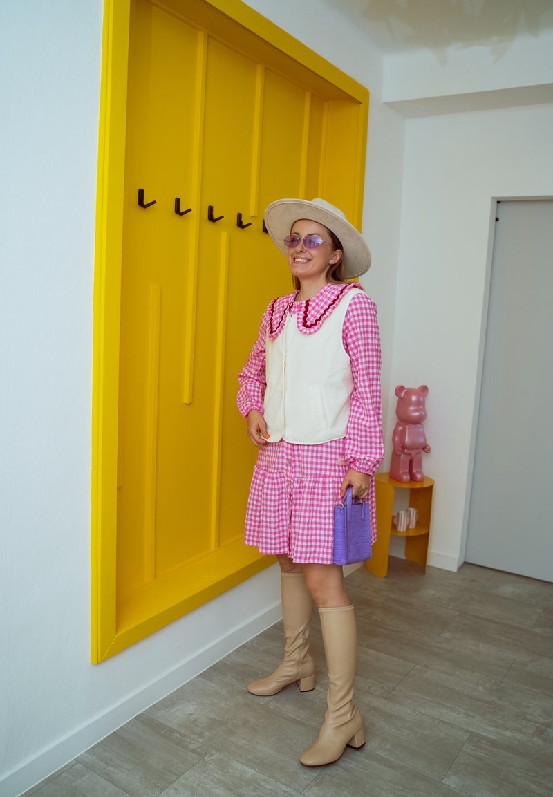 Vestito rosa a quadri con orlo arricciato, gilet bianco con colletto, stivali beige fino al ginocchio, borsa viola e cappello a tesa larga, su sfondo di parete gialla.