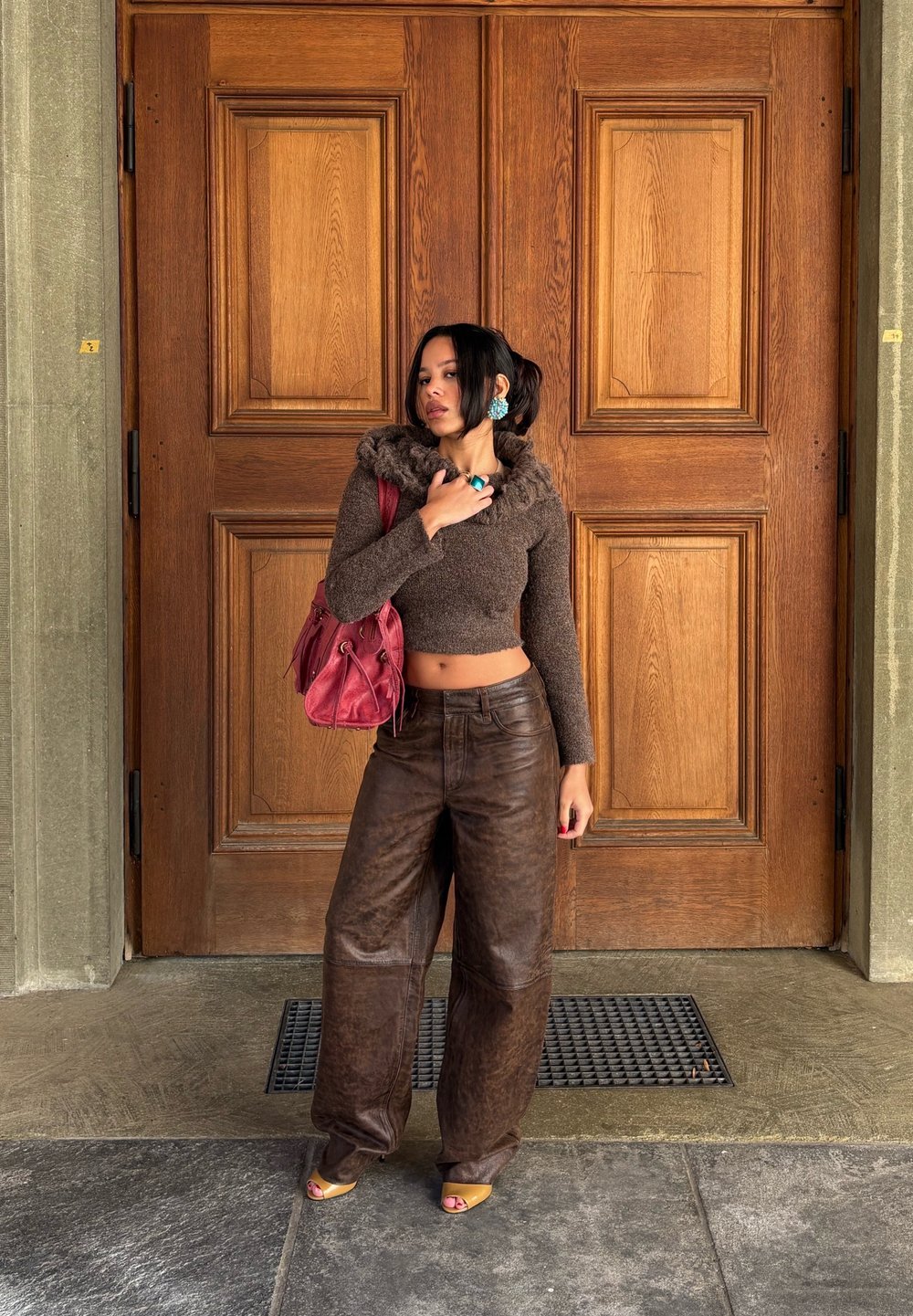 Woman standing in front of large wooden double doors wearing a brown cropped sweater, baggy leather trousers, yellow sandals, and a red handbag.