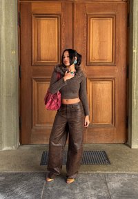 Woman standing in front of large wooden double doors wearing a brown cropped sweater, baggy leather trousers, yellow sandals, and a red handbag.