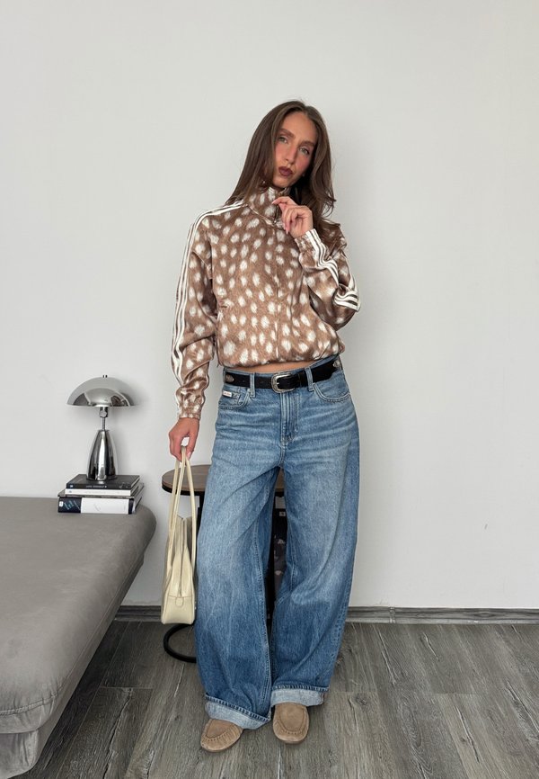 Mujer que lleva una chaqueta con cremallera y manchas, vaqueros azules anchos y mocasines, sosteniendo un bolso claro, de pie en una habitación moderna con una lámpara y libros.