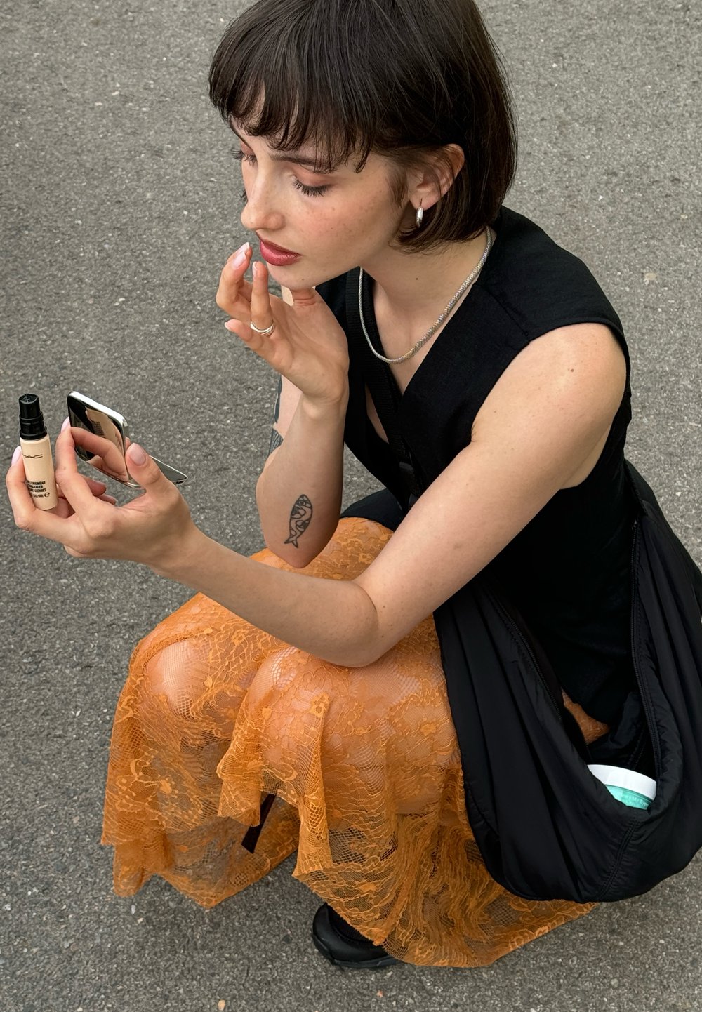 Svart ärmlös topp, orange spets midi kjol, applicerar produkt från en liten flaska, håller en spegel. Asfaltbakgrund, minimala accessoarer.