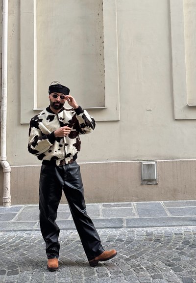 Chaqueta de piel sintética con un patrón de vaca en blanco y negro, pantalones de cuero negro, botas marrones y una boina negra con gafas de sol. De pie en una calle de adoquines.