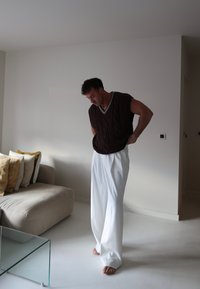 Rjava pletena brezrokavna majica z V-izrezom, v kombinaciji z belimi širokimi hlačami. Prikazuje teksturiran, sproščen videz v minimalističnem okolju.