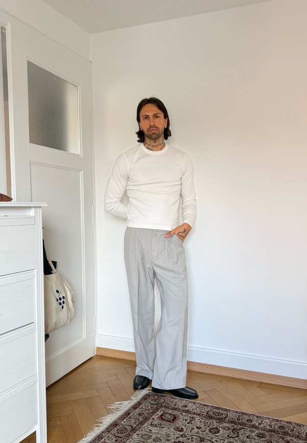 Homme avec des tatouages portant une chemise blanche à manches longues, un pantalon large gris clair et des chaussures noires, debout dans une pièce minimaliste.
