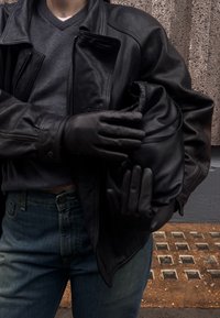 Mustast nahast jakk ja kindad, mida kannab isik, kes hoiab musta, kogutud kotti, kombineeritud siniste teksadega. Taustal on tekstuuriga pinnad.