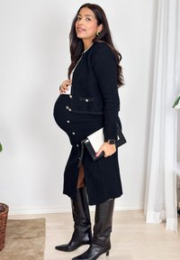Femme enceinte debout à l'intérieur, portant un cardigan noir, une jupe boutonnée, des bottes montantes, tenant une pochette noire et blanche.