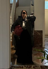 Μαύρη δερμάτινη τζάκετ πάνω από ένα γραφικό t-shirt, μαύρη φούστα, και κρατώντας μια κόκκινη τσάντα. Ο φόντος περιλαμβάνει σκάλες και εσωτερικά φυτά.