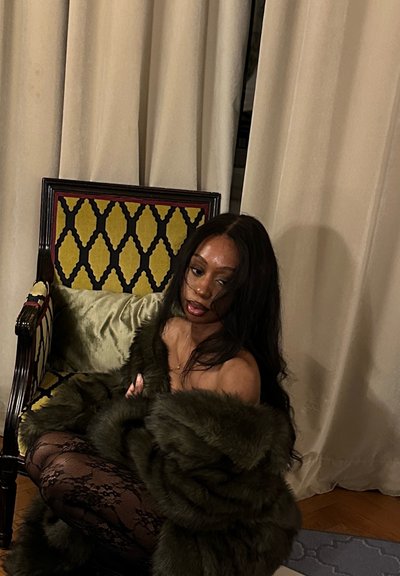 Mujer de cabello largo sentada en el suelo junto a una silla con estampado, lleva un abrigo de piel oscuro y medias de encaje negras frente a un fondo de cortinas beige.