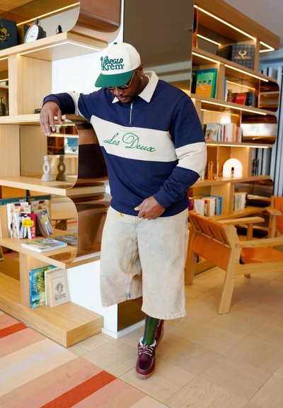 Pulóver azul marino y crema con bloques de color y logo "Les Deux", combinado con pantalones cortos de mezclilla claros y zapatos burdeos con cordones. Gorra blanca con detalles verdes.