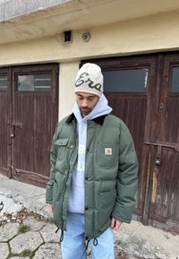 Doudoune vert olive avec fermeture éclair et boutons, poches avant et col marron foncé ; portée sur un sweat à capuche gris et un jean bleu.