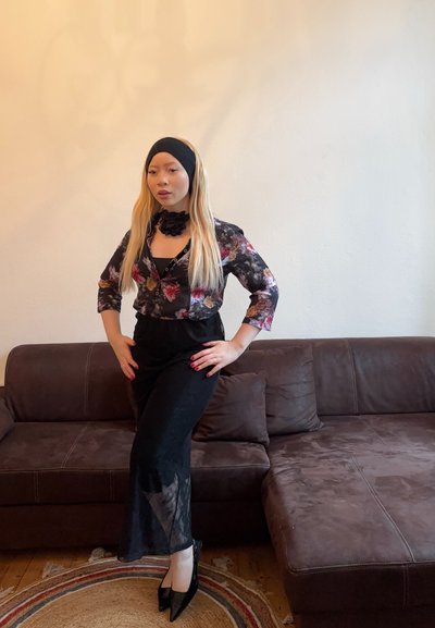 Blusa negra con estampado floral y mangas de tres cuartos, falda negra con paneles transparentes, diadema negra y zapatos de tacón alto, posando en un sofá marrón.