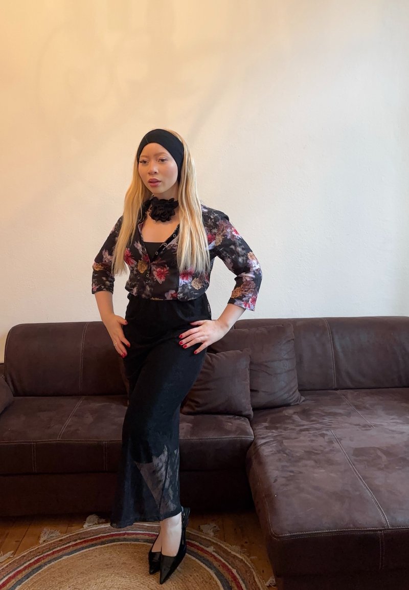 Black floral blouse with three-quarter sleeves, black skirt with sheer panels, black headband, and high-heeled shoes, posed on a brown sofa.