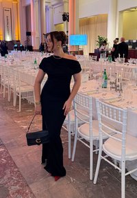 Femme en robe noire asymétrique et talons rouges tenant un sac à main noir, debout près de tables de banquet blanches dressées avec de la verrerie et des bougies.
