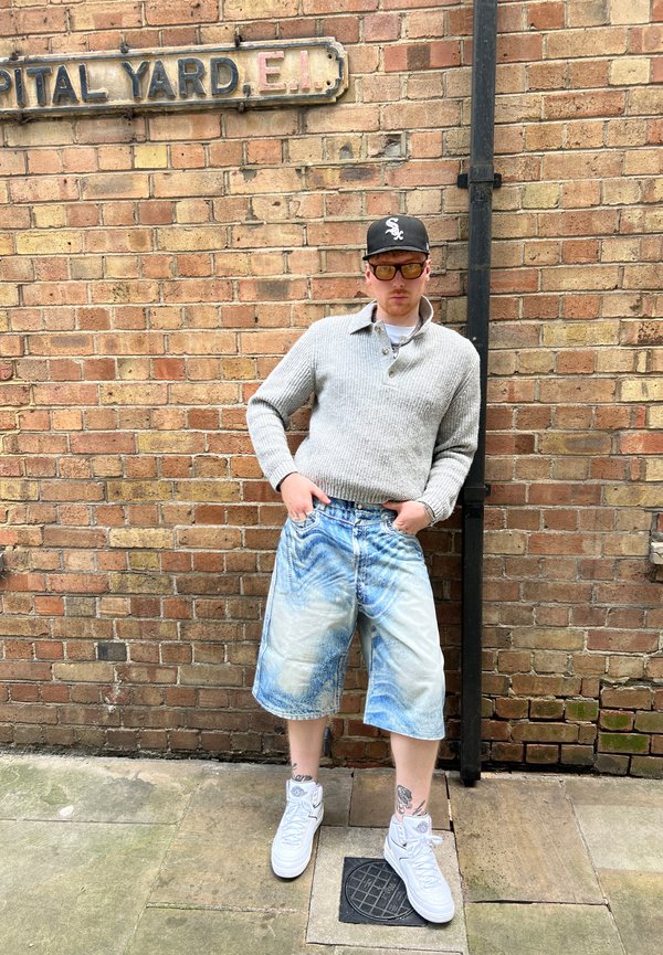 Suéter de punto gris claro, pantalones cortos de mezclilla azul de gran tamaño con un patrón desteñido y zapatillas altas blancas frente a un fondo de pared de ladrillo.