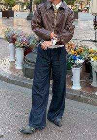 Giacca di pelle marrone con cuciture bianche a contrasto, camicia bianca, pantaloni larghi in denim scuro e scarpe nere, in piedi vicino a composizioni floreali.