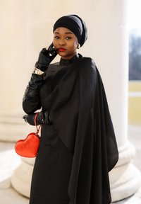 Γυναίκα με μαύρο καλυμμα κεφαλής και γάντια, φοράει χρυσά κοσμήματα, κρατά κόκκινη τσάντα σε σχήμα καρδιάς, ποζάροντας με το χέρι κοντά στο πρόσωπο σε φωτεινό φόντο.