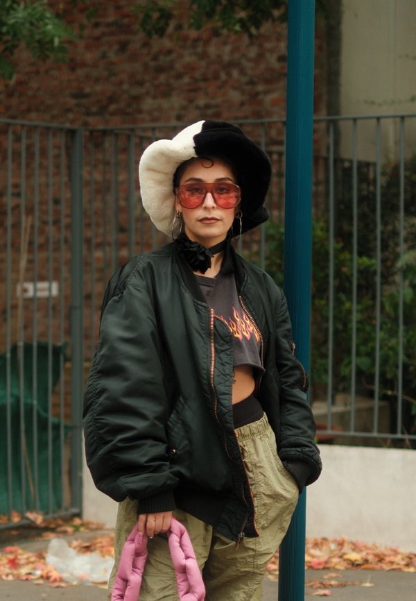 Person iført sort og hvid hat, oversized mørk jakke, crop top med flammeprint, beige bukser, lyserød flettet taske og store solbriller med lyserøde brilleglas.