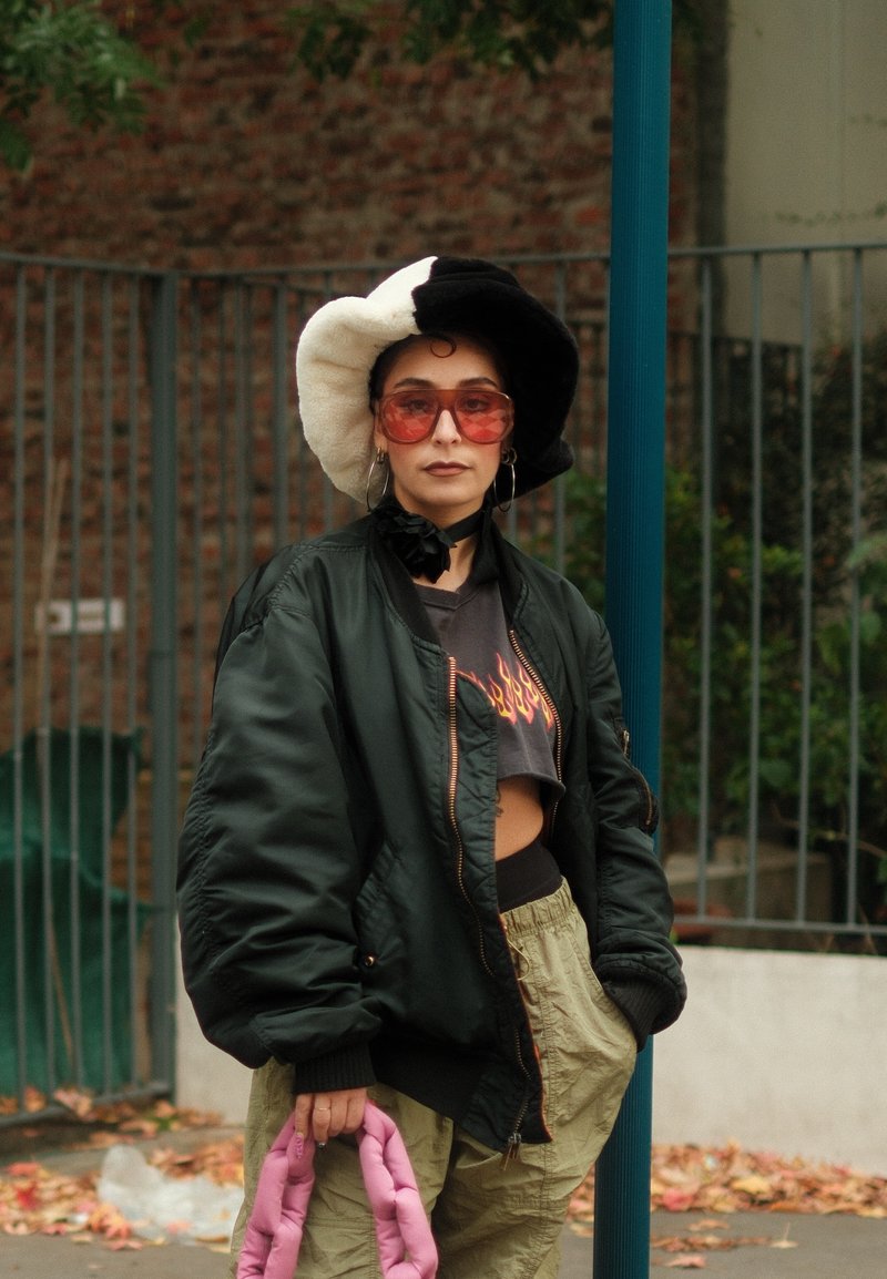 Person trägt einen schwarz-weißen Hut, eine übergroße dunkle Jacke, ein Cropped-Top mit Flammenmuster, beige Hosen, eine pink geflochtene Tasche und große, pink getönte Sonnenbrillen.