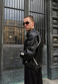 Muškarac u crnoj kožnoj jakni i sunglassesima stoji pored ukrašenih metalnih vrata s prozorima rešetki, gledajući u stranu.
