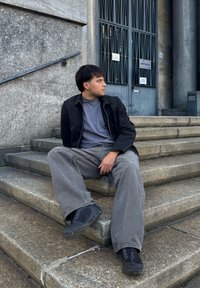 Čierna zipová bunda cez šedé tričko, šedé široké nohavice a čierne topánky, sedí na kamenných schodoch pri vstupe do budovy.