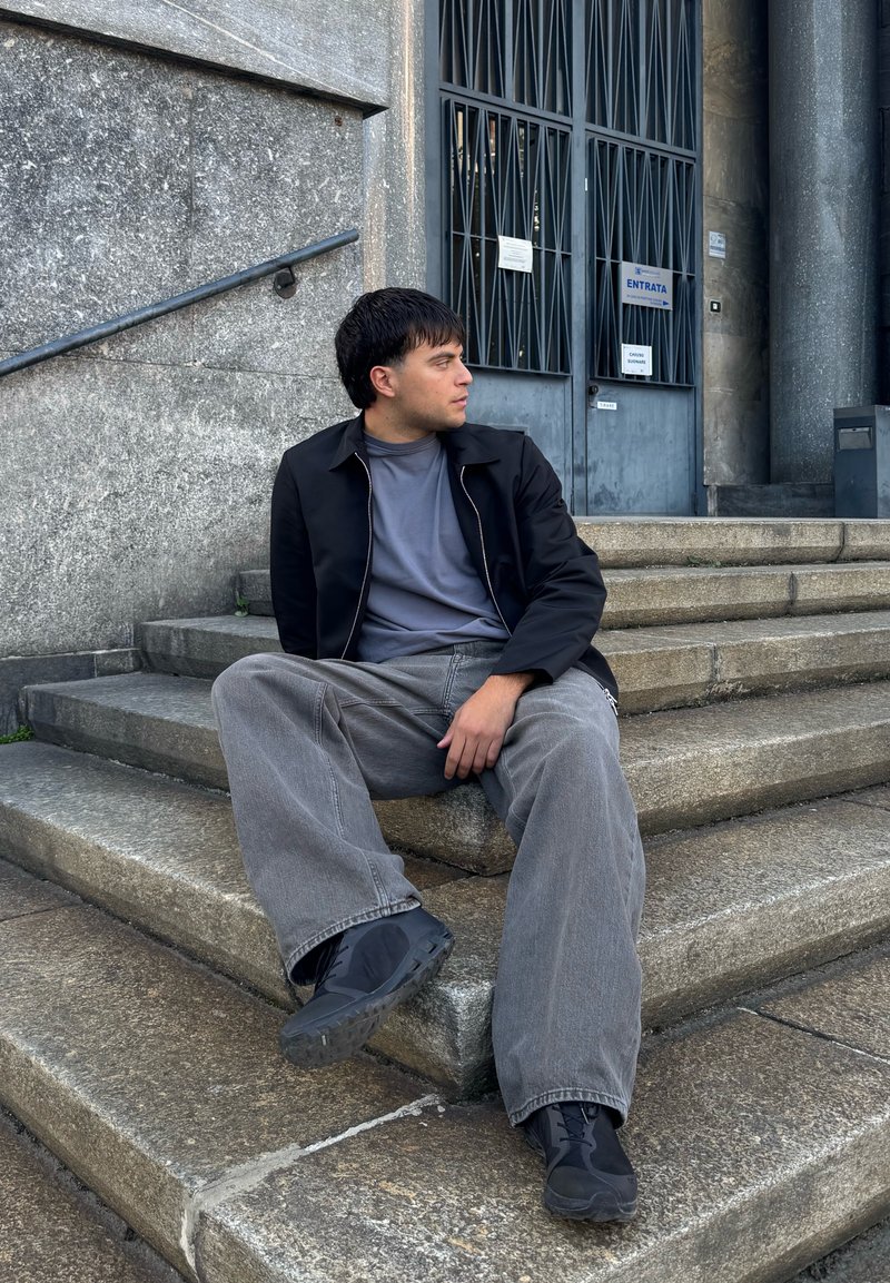 Veste à zip noire sur un t-shirt gris, pantalon large gris et chaussures noires, assis sur des marches en pierre près de l'entrée d'un bâtiment.