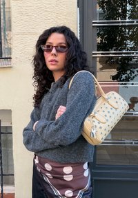 Femme aux cheveux bouclés et foncés portant des lunettes de soleil, un pull gris et une jupe à pois, portant un sac à main beige avec des œillets en métal.