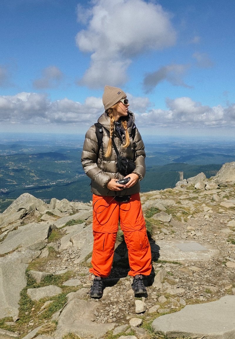 Une personne portant une veste en duvet grise et un pantalon cargo orange se tient sur un terrain rocheux, tenant un appareil, avec des cheveux tressés et des lunettes de soleil.