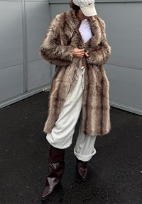 Personne portant un manteau en fausse fourrure beige, un crop top blanc, un pantalon de survêtement gris clair, des bottes marron foncé et une casquette Nike beige, debout sur un trottoir noir.