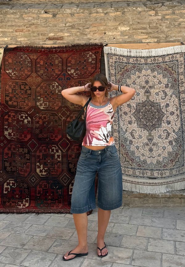 Joven mujer con gafas de sol y ropa informal está entre dos alfombras colgadas con patrones, sobre un suelo de piedra con una pared de ladrillos de fondo.