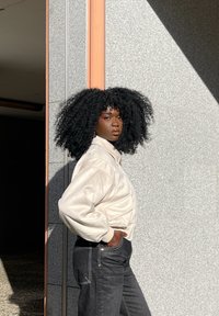 Jachetă bej pal, matlasată, cu guler înalt, purtată peste blugi negri din denim. Fundalul prezintă o suprafață gri texturată și lumina soarelui.