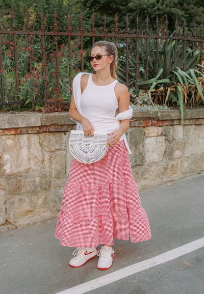 Top blanco sin mangas, falda de cuadros rojos con volantes en capas, bolso blanco en forma de media luna, zapatillas blancas con acentos rojos, gafas de sol; entorno al aire libre.
