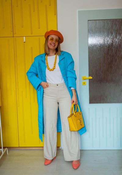 Mujer con boina naranja, gafas de sol amarillas, abrigo azul, camisa blanca, pantalones beige, zapatos naranjas, collar amarillo, sosteniendo un bolso amarillo de pie en interiores.